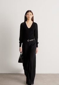 Sieviete ar gariem tumšiem matiem tērpusies pilnībā melnā apģērbā ar V-veida izgriezuma topu, platu kāju biksēm, jostu un nēsā melnu rokassomu, stāvot pret vienkāršu fona sienu.