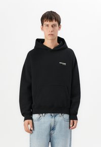 Haut à capuche noir avec une poche kangourou, des poignets et un bas en côte, présentant un petit logo blanc et doré sur la poitrine gauche. Texture de tissu douce.