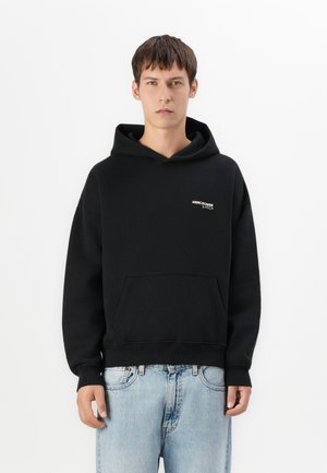 Haut à capuche noir avec une poche kangourou, des poignets et un bas en côte, présentant un petit logo blanc et doré sur la poitrine gauche. Texture de tissu douce.