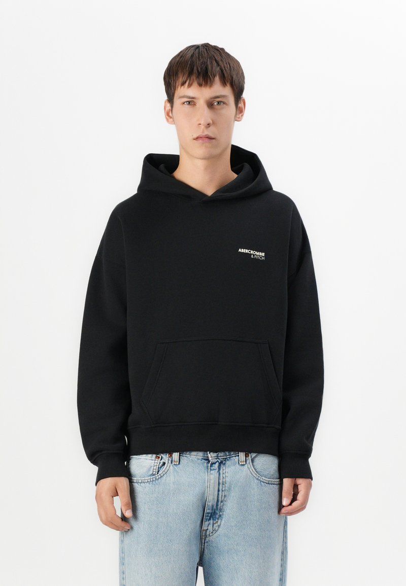 Haut à capuche noir avec une poche kangourou, des poignets et un bas en côte, présentant un petit logo blanc et doré sur la poitrine gauche. Texture de tissu douce.