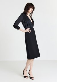 Schwarzes Satin-Kleid mit tiefem V-Ausschnitt und langen Ärmeln, mit tailliertem Schnitt und A-Linien-Rock. Kombiniert mit schwarzen High-Heels.