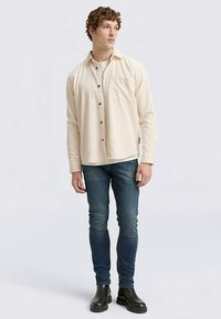 Camicia color crema con bottoni e taschino, abbinata a jeans blu scuro e stivaletti neri alla caviglia. Il tessuto appare strutturato. Vestibilità casual.