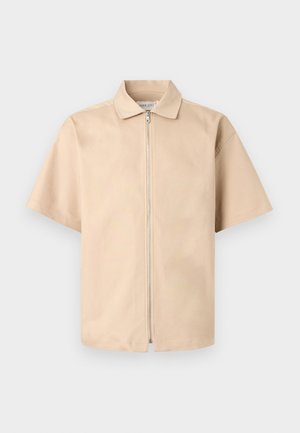 Beige kortermet skjorte med krage og glidelås foran, vist mot en ensfarget lys bakgrunn.