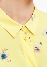 Gros plan d'une chemise jaune à col avec deux boutons jaunes et des imprimés floraux subtils en bleu, rose et noir près du col et de la poitrine.