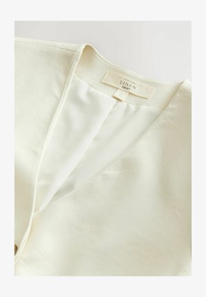 Krēmo krāsas linu tops ar V veida kakla izgriezumu, kuram ir gluda iekšējā oderēšana un etiķete, kas norāda auduma sastāvu. Vienkāršs pogu aizdare.