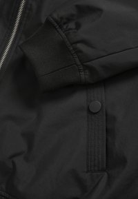 Črno bomber jakno z zadrgo, rebrastimi manšetami in detajlom s pritisnim gumbom, izdelano iz gladkega, lahkega blaga.