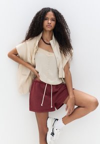 Beige knapplöst väst över en ljus camisole med spetsdetaljer, tillsammans med maroon shorts och vita sneakers med svarta detaljer.