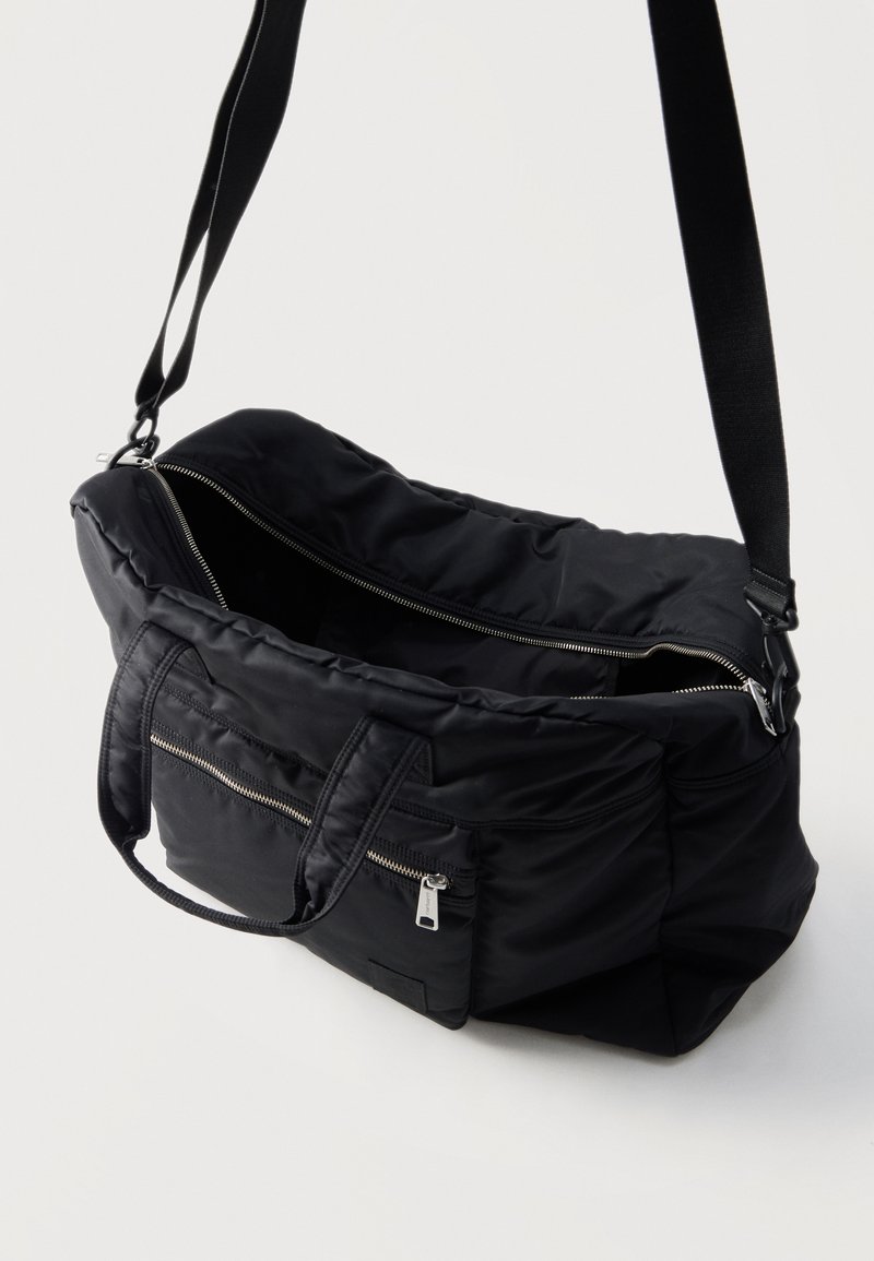 Zwarte nylon duffeltas met open hoofdrits, voorvak met rits, twee handvatten en een verstelbare schouderriem op een lichte achtergrond.