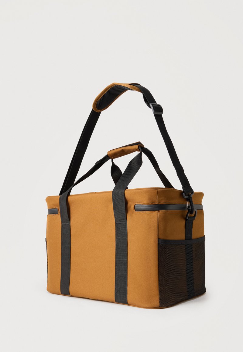 Bolsa de lona rectangular marrón con asas negras, correa de hombro ajustable, bolsillo lateral de malla y cierre superior con cremallera sobre un fondo liso.