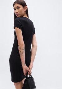 Μαύρο εφαρμοστό φόρεμα με κοντά μανίκια, λείο υφή. Το μοντέλο κρατά μια μικρή μαύρη τσάντα· ορατά τατουάζ στο αριστερό μπράτσο.