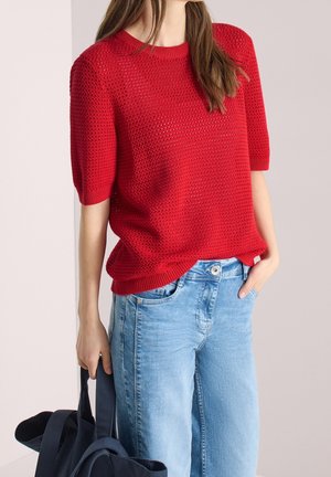 Personne portant un pull en maille rouge à manches courtes et un jean bleu clair, tenant un sac bleu foncé dans une main et l'autre main dans une poche.