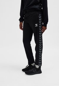 Person trägt schwarze Trainingshose mit weißem Hummel-Logo und Chevron-Streifen an den Seiten, kombiniert mit schwarzen Sneakern und weißen Socken.