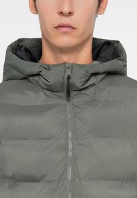 Γκρι φουσκωτό μπουφάν με κουκούλα, διαθέτει πλήρη φερμουάρ και ραφτό υλικό, τονίζοντας μια λεία εξωτερική επιφάνεια και μαύρη εσωτερική επένδυση.