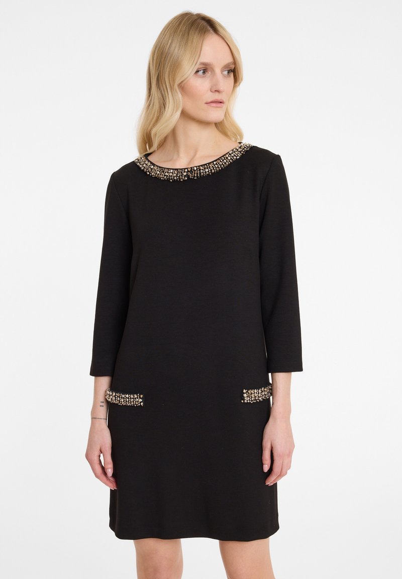 Femme blonde portant une robe noire à mi-mollet avec un col en perles et des poches, debout contre un fond blanc, regardant vers la droite.