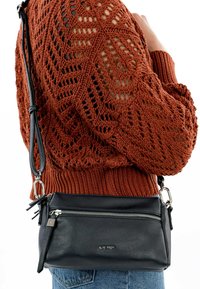 Borsa a tracolla nera in pelle con chiusura a zip, dotata di una tracolla staccabile e accessori in metallo argentato, esposta su un maglione in rust texturizzato.