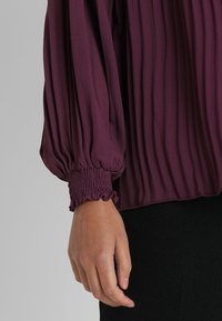 Handen vilar vid sidan iklädd en långärmad plommonfärgad blus med samlade elastiska manschetter och plisserade tygdetaljer.