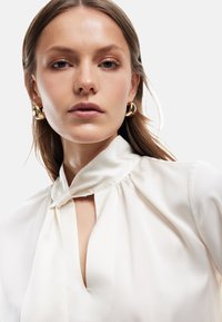 Weiße Satin-Bluse mit hohem Kragen und Schlüssellochdetail. Verfügt über sanfte Drapierung und eine glatte Textur, ergänzt durch kleine goldene Creolen.