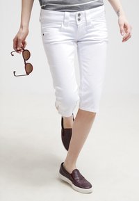 Valkoiset lyhyet housut ovat slim fit -malliset, niissä on kahden napin sulkumekanismi ja lievät sivuhalkiot. Yhdistetty tummiin liukuhyppäreihin ja aurinkolaseihin.