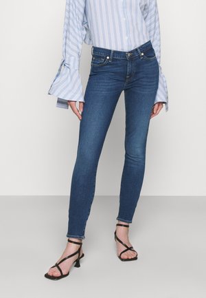 Femme portant un jean skinny bleu, un chemisier à rayures bleues et blanches avec des manches évasées, et des sandales à talons noires à brides, sur un fond uni.