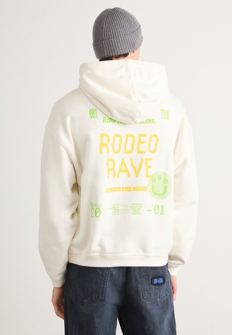 Krēmveida kapuce ar lielu dzeltenu tekstu "RODEO RAVE", zaļiem akcentiem un jautrām grafikām. Apvienots ar tumši zilām džinsām. Ribota pelēka cepure.