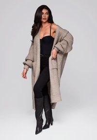 Langer gestrickter Cardigan in Beige, über einem schwarzen Bodysuit und schwarzen Skinny Jeans getragen. Das Modell trägt schwarze kniehohe Stiefel.