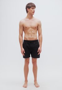 Μαύρα μαγιό σορτς με ρυθμιζόμενο κορδόνι στη μέση, κατασκευασμένα από ελαφρύ υλικό, παρουσιάζονται σε όρθιο αρσενικό μοντέλο.