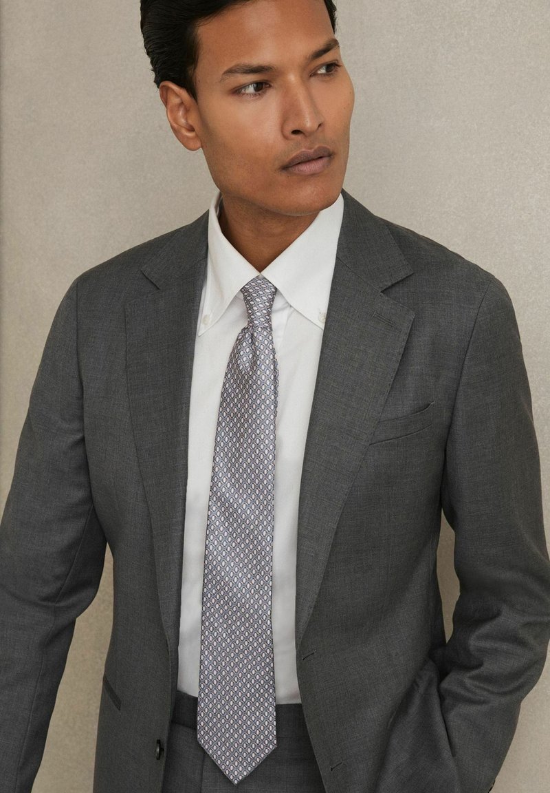 Costume gris avec une texture subtile, chemise blanche et cravate légère violette à motifs. Détails notables incluant le revers et un accessoire de poche.