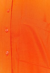 Pomarańczowa koszula zapinana na guziki, wykonana z lekkiego materiału, z kieszenią na lewej piersi, widocznym przeszyciem oraz pięcioma pasującymi guzikami na listwie.