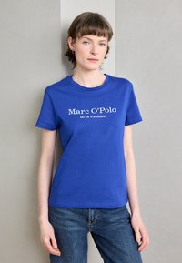 Modrá bavlněná tričko s krátkým rukávem, na přední straně s bílým textem "Marc O'Polo EST. V STOCKHOLMU" umístěným uprostřed. V kombinaci s džínovými kalhotami.