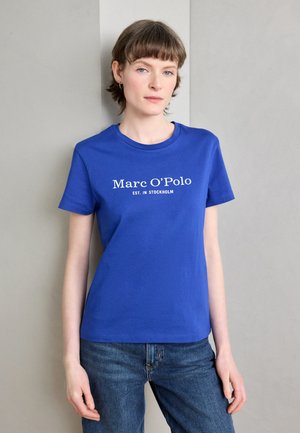 Sinine puuvillane T-särk lühikeste varrukatega, millel on ees keskelt valge tekst "Marc O'Polo EST. IN STOCKHOLM". Kombineeritud teksapükstega.