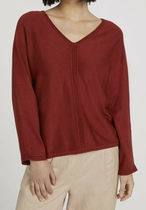 Pull en tricot couleur rouille avec un col en V, des manches longues et un discret détail côtelé sur le devant. Texture lisse, coupe loose.