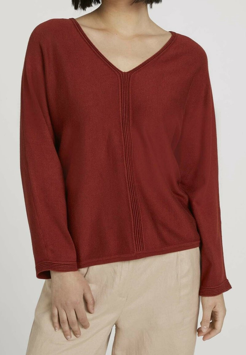 Pull en tricot couleur rouille avec un col en V, des manches longues et un discret détail côtelé sur le devant. Texture lisse, coupe loose.