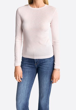 Woman wearing a fitted long-sleeve pink and white striped shirt paired with blue jeans, standing against a plain light background.