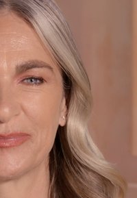 Zbliżenie twarzy kobiety z jasnobrązowymi włosami, subtelnymi falami, gładką skórą i delikatnym kolorem ust. Przekłuty ucho z małym kolczykiem.