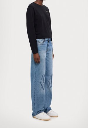 Personne portant un t-shirt noir à manches longues, un jean large bleu clair avec de grands logos "A|X" sur les cuisses, et des baskets blanches à semelles en gomme.