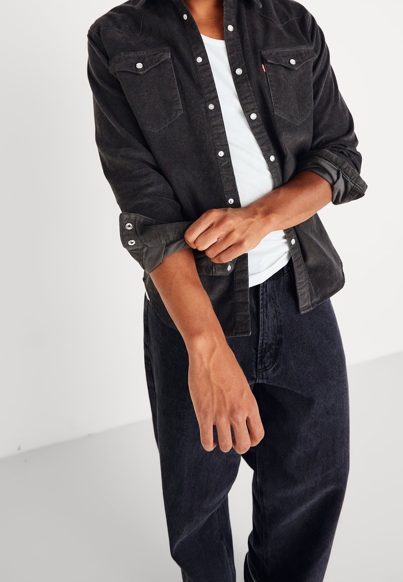 Personne retroussant la manche d'une chemise en velours côtelé noire à boutons portée sur un t-shirt blanc et un jean foncé sur un fond uni.