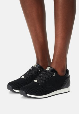 Gambe con sneakers in maglia nera con lacci neri, targhette con logo argentato e suole bianche, su uno sfondo chiaro e uniforme.
