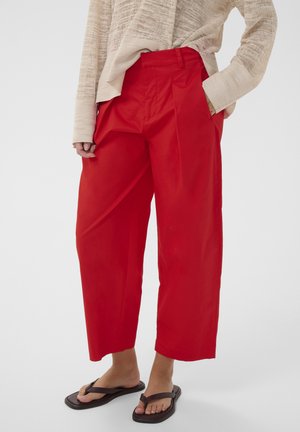 Femme portant un pantalon large rouge vif raccourci, un haut beige texturé à manches longues et des tongs noires, debout avec une main dans la poche.