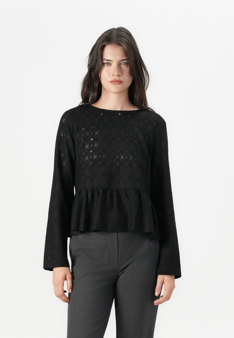 Top peplum nero realizzato in tessuto strutturato con maniche lunghe e collo rotondo, caratterizzato da un sottile motivo geometrico. Abbinato a pantaloni grigi.