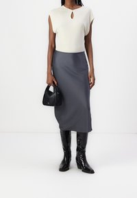 Kobieta w białej bluzce bez rękawów, szarej spódnicy midi, czarnych lakierowanych botkach do kolan i trzymająca małą czarną torebkę.