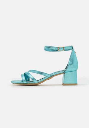 Sandale turquoise métallisée à bout ouvert avec talon bloc, brides croisées et boucle réglable à la cheville sur fond blanc.