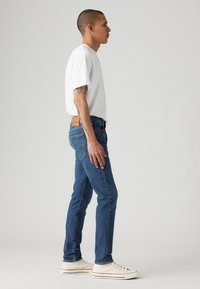 Blaue Jeans im Slim-Fit-Stil, kombiniert mit einem kurzärmeligen weißen T-Shirt und weißen Sneakers, von der Seite betrachtet.