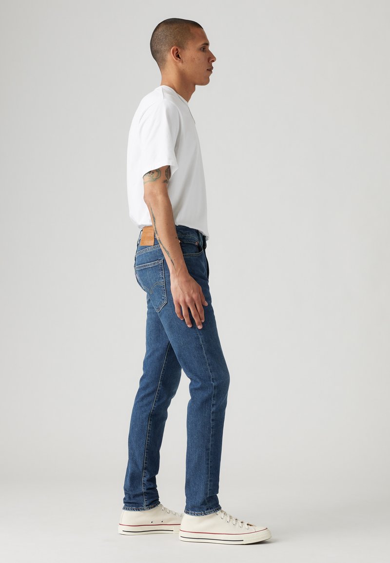 Blauwe denim jeans met een slim fit, gecombineerd met een wit t-shirt met korte mouwen en witte sneakers, bekeken vanuit de zijkant.