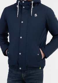 Homme portant une veste à capuche bleu marine avec des cordons blancs, des boutons-pression, les mains dans les poches avant, associée à un jean bleu foncé.