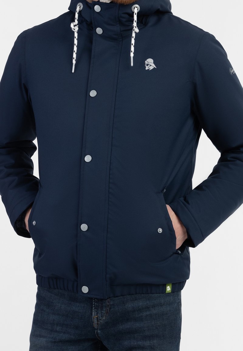 Homme portant une veste à capuche bleu marine avec des cordons blancs, des boutons-pression, les mains dans les poches avant, associée à un jean bleu foncé.