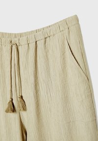 Pantalon beige texturé avec une ceinture élastique et des cordons à pompons, avec une poche latérale droite.