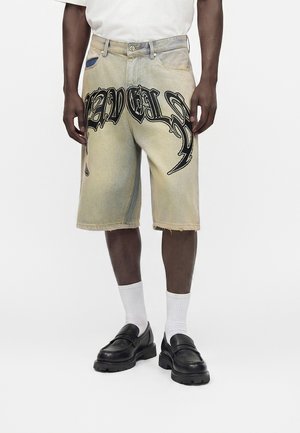 Person, die beige kniehohe Jeansshorts mit schwarzem gotischem Textdesign, weißen Socken und schwarzen Mokassins trägt, steht vor einem weißen Hintergrund.