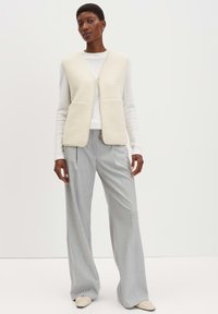 Cremfarbener Fleece-Weste mit einem Reißverschluss auf der Vorderseite, getragen über einem weißen Langarmshirt. Kombiniert mit hellgrauen, weit geschnittenen Hosen und schlichten Schuhen.