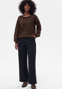 Kobieta o kręconych włosach, ubrana w brązowy sweter we wzory, czarne spodnie o szerokich nogawkach oraz brązowe botki, stoi z jedną ręką w kieszeni.