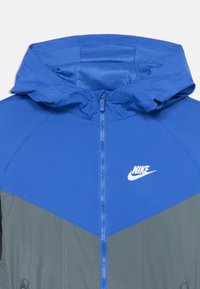 Nike striukė su mėlynu kapišonu ir viršutine kūno dalimi, pilkos spalvos apatine dalimi, užtrauktuku ir logotipu ant krūtinės. Glotni tekstūra su sportiniu dizainu.